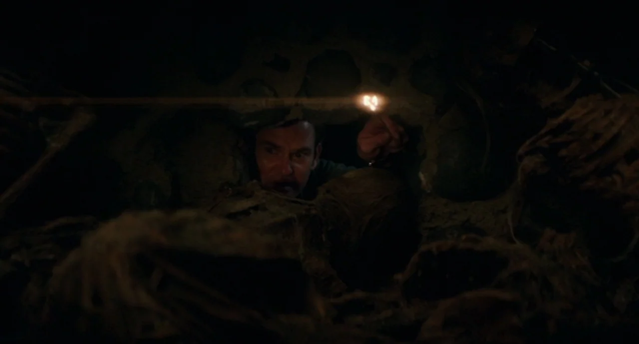 Ouija: Origin of Evil (2016) movie still: medium — A man peers through a narrow, dark opening surrounded by mounds of earth. He holds a single…