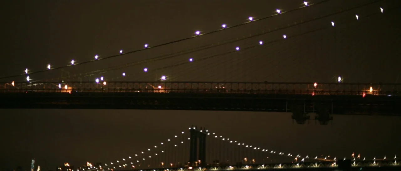 NY84 (2016) movie still: establishing — Two large steel suspension bridges are visible at night against a dark backdrop. The structures are outlined…