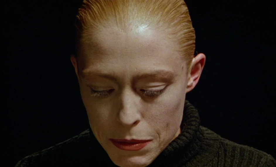Wittgenstein (1993) movie still: closeup — A person is centered against an undifferentiated black background. The figure gazes downwards with eyes…