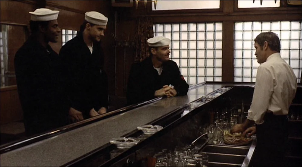 The Last Detail (1973) movie still: medium — A dimly lit room features wood-paneled walls and a long bar with glass bricks. Three sailors wearing…