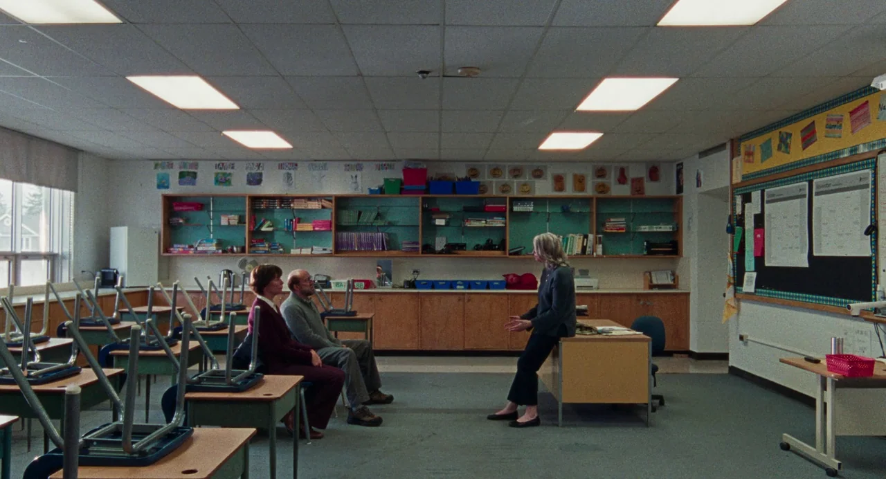 Dream Scenario (2023) movie still: wide — An elementary classroom with upside-down chairs on desks and shelves filled with books. A woman stands by…