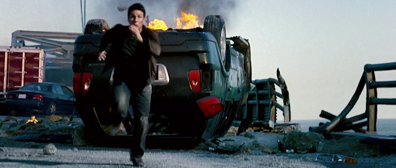 Down With Love (2003) movie still: fullbody — An asphalt road surface on a bridge features a flipped, burning SUV and debris. A man runs towards the…