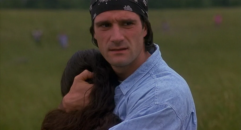 Exotica (1994) movie still: medium — An open grassy field stretches into the blurred distance under an overcast sky. A man in a bandana holds a…