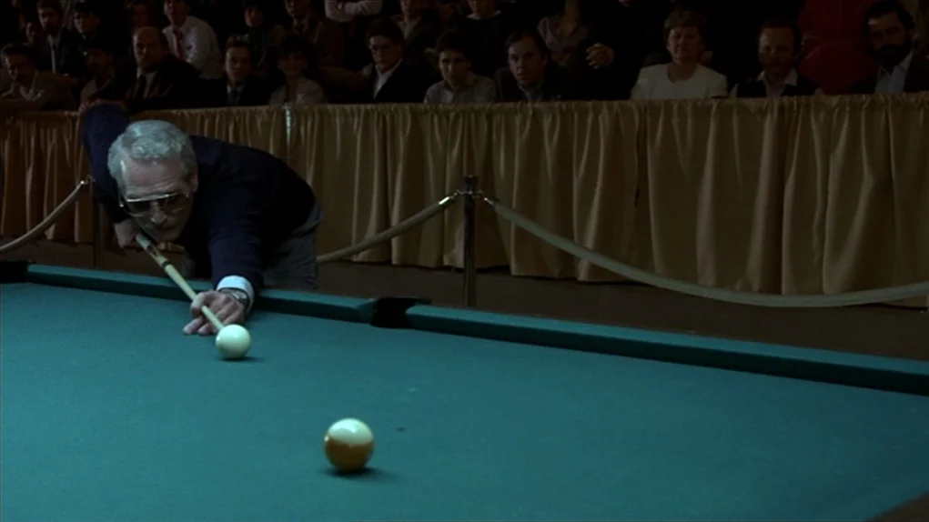 The Colour of Money (1986) movie still: medium — A green-felt pool table sits in front of a seated audience in a dim room. An older man in aviator…