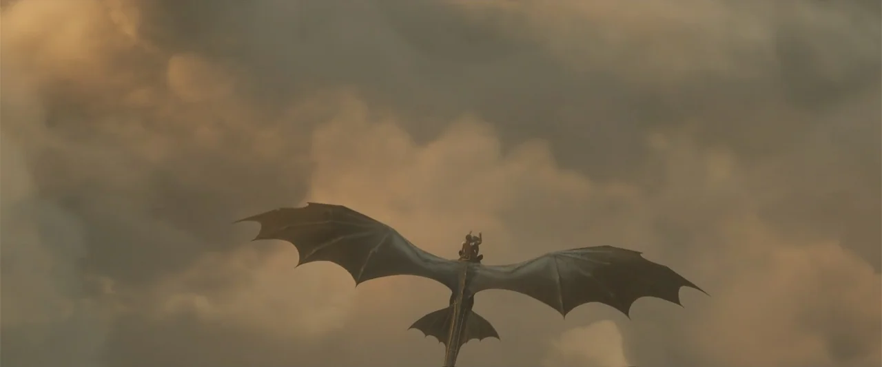How to Train Your Dragon (2025) movie still: wide — A massive winged dragon silhouette drifts through thick, layered golden clouds at sunset. A…