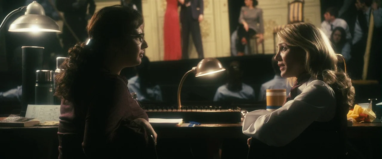 The Box (2009) movie still: medium — A dimly lit production booth contains a long wooden desk with sound equipment. Two women sit facing each other…