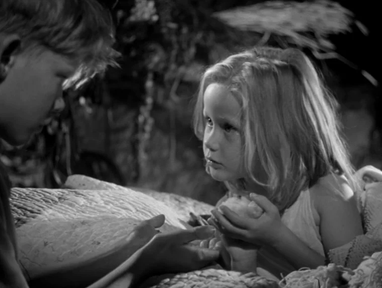 Forbidden Games (1952) movie still: medium — A dimly lit room at night features a textured white blanket in the foreground. A young girl and a young…