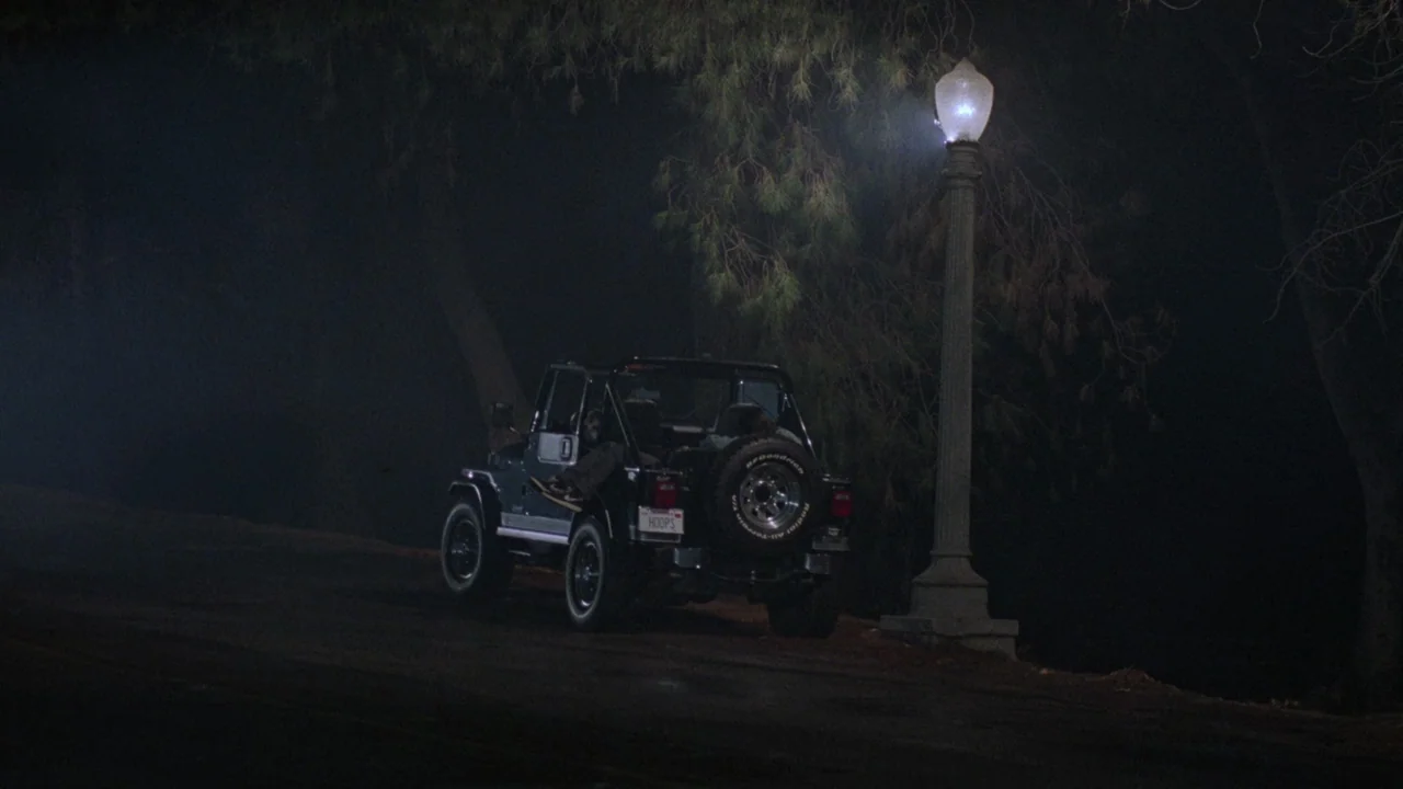 Society (1989) movie still: wide — A dark, paved road winds through trees at night under a single lamp. An off-road jeep is parked facing away from…