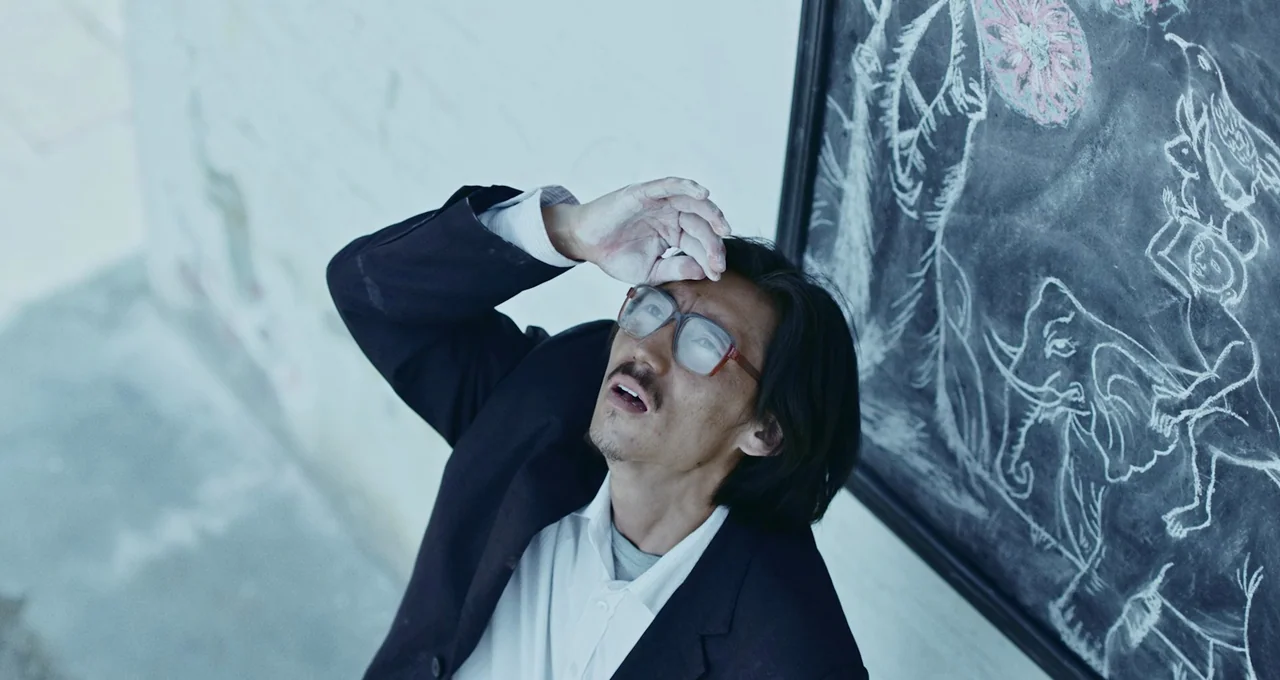 Balloon (2019) movie still: medium — A man stands indoors next to a large chalkboard featuring white chalk animal drawings. The man gazes upward…