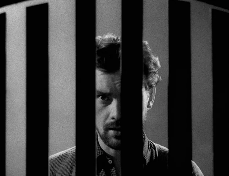 Prison (1949) movie still: closeup — A man stands behind heavy iron bars inside a dark stone room. The prisoner peers out from between the vertical…