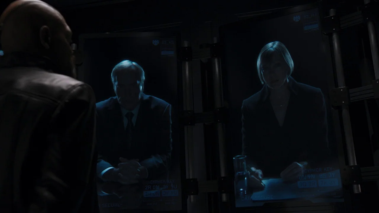 The Avengers (2012) movie still: medium — A dark, high-tech interior features two large vertical monitors displaying video feeds of two individuals.…