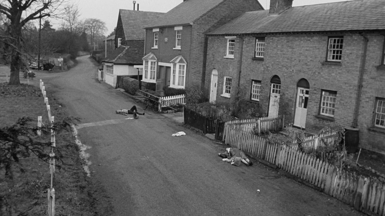 Village of the Damned (1960) movie still: wide — A quiet English village road passes by a row of small brick houses. Two motionless figures lie…