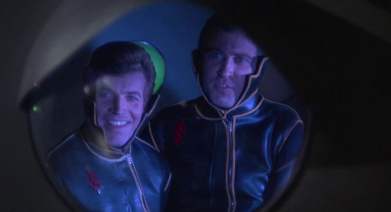 Planet of the Vampires (1965) movie still: medium — Two men in futuristic uniforms stand inside a dim, blue-lit cockpit or observation area. They…