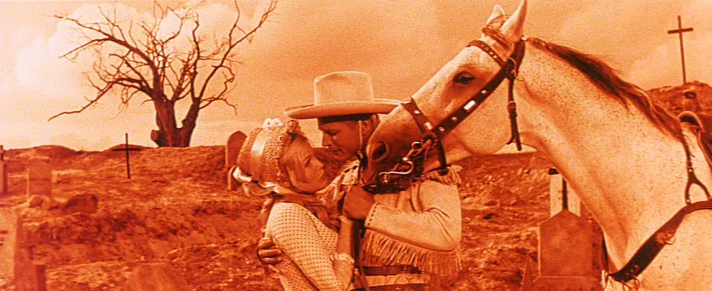 Lemonade Joe (1964) movie still: medium — A desolate cemetery contains wooden grave markers and a single leafless, barren tree. A man and a woman in…