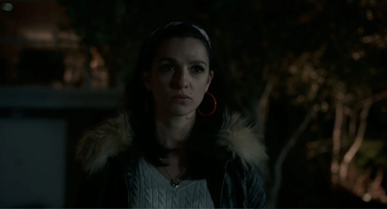 Back to Black (2024) movie still: medium — A woman stands in a dark residential driveway at night with foliage behind her. She faces the camera…