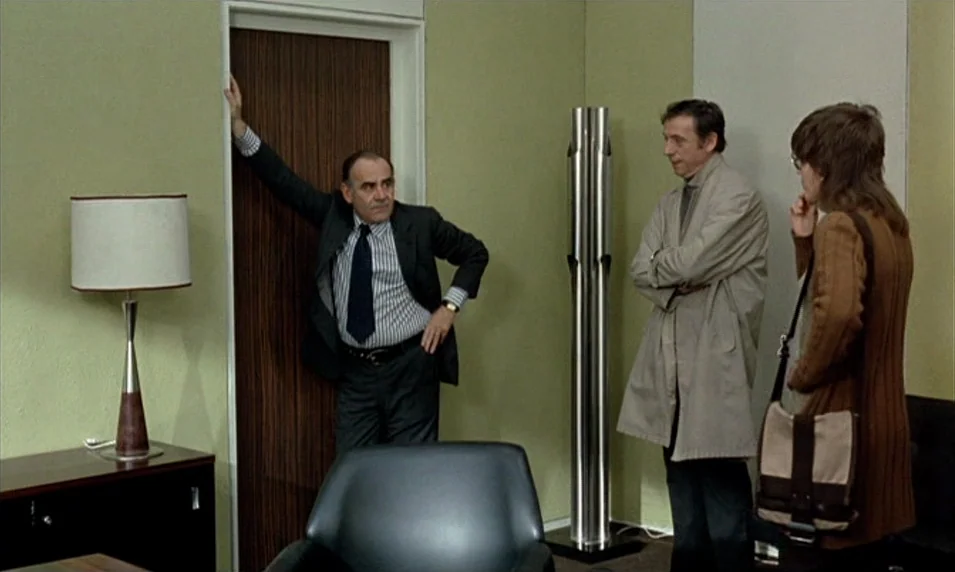 Tout Va Bien (1972) movie still: medium — A 1970s room with light olive walls and a tall modern metal sculpture. Three people interact in the space,…