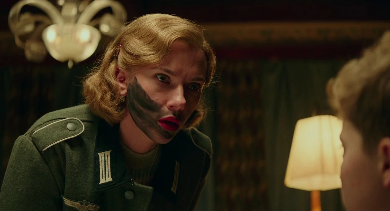 Jojo Rabbit (2019) movie still: medium — A dimly lit room features a woman wearing a military jacket and face paint. She leans forward towards a…