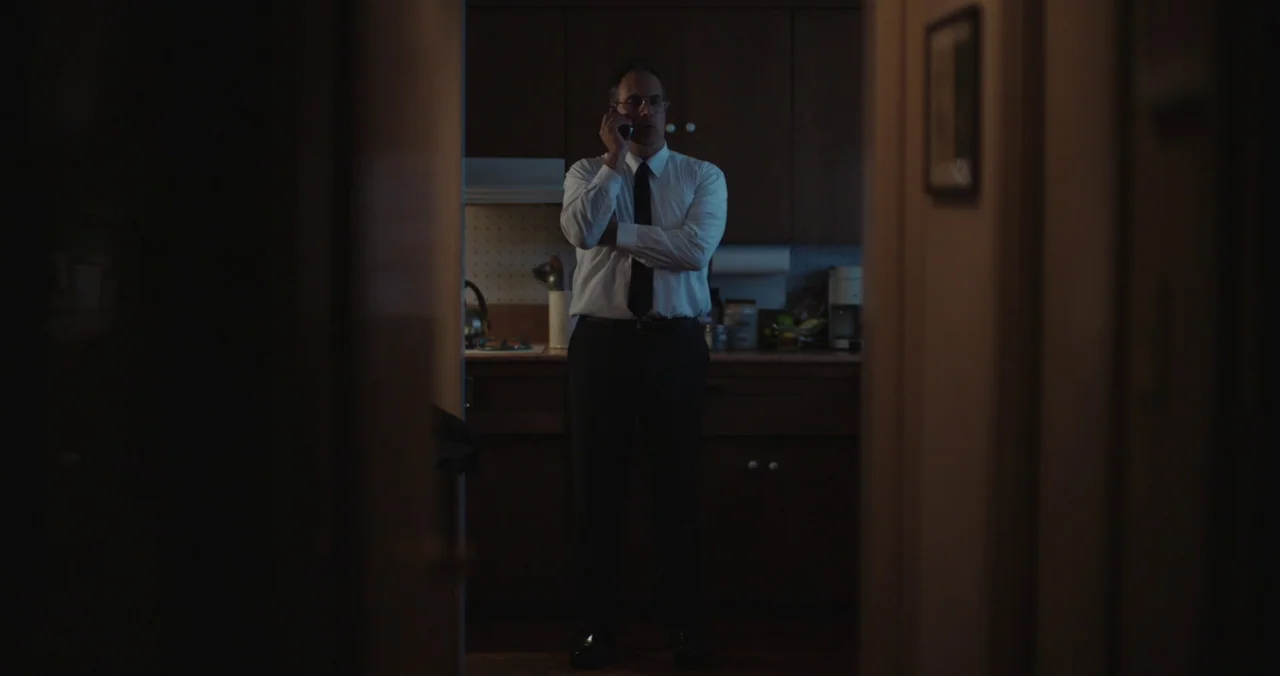 Violet (2021) (2021) movie still: medium — A dimly lit room with dark wood cabinetry and a countertop visible in back. A man wearing a dress shirt…
