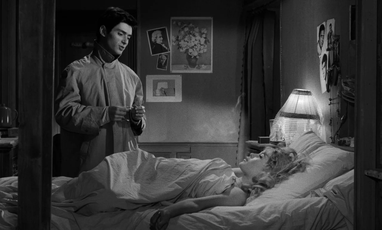 La Verite (1960) movie still: medium — A dimly lit room with walls decorated with photographs and a lamp. A man in a coat watches a woman resting in…