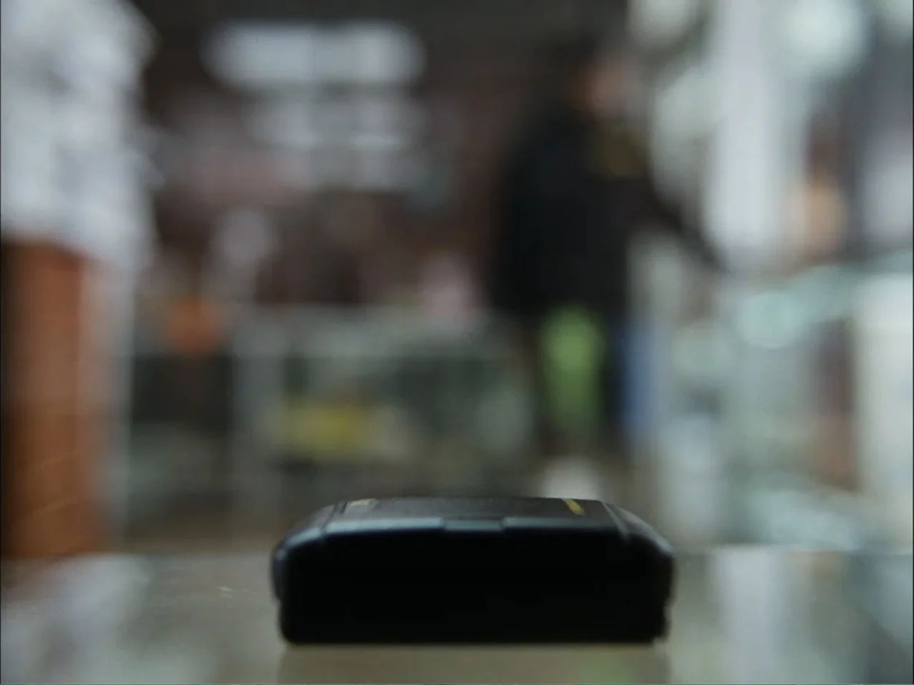 As We Speak: Rap Music on Trial (2024) movie still: closeup — A dark, rectangular object sits on a reflective surface inside a blurred shop. The…