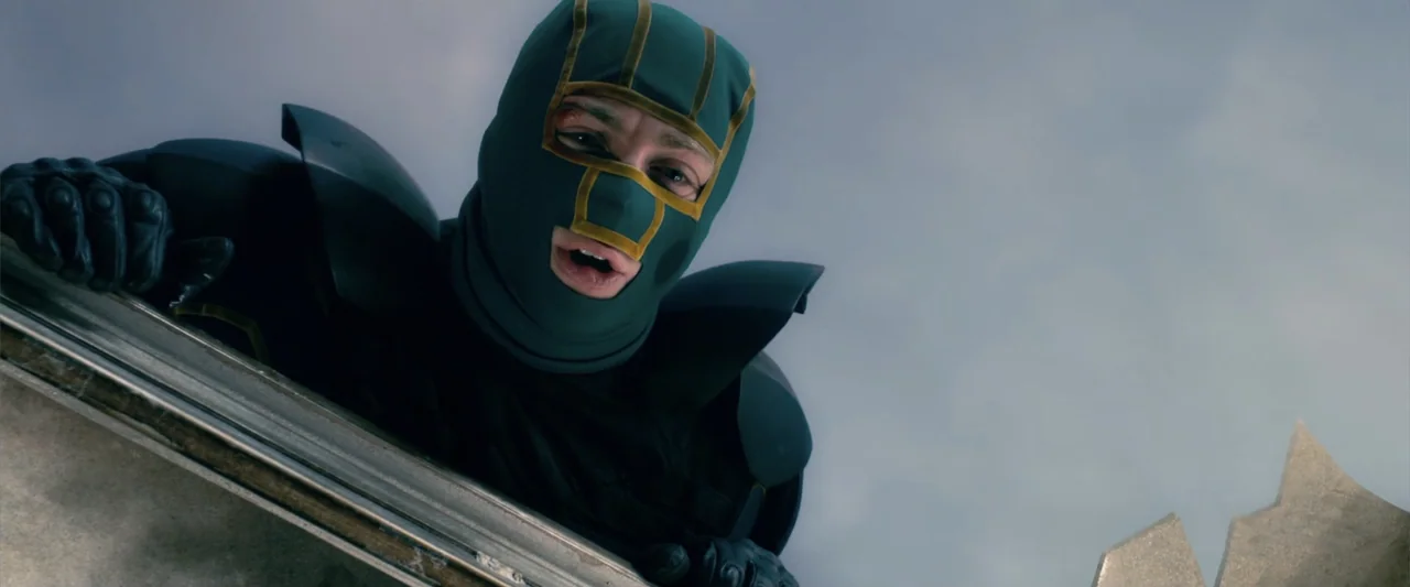Kick-Ass 2 (2013) movie still: medium — A rooftop edge is visible against a cloudy grey sky. A man in a tactical mask leans forward looking down at…