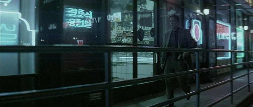Heat (1995) movie still: fullbody — A sidewalk runs past the illuminated glass windows of a convenience store at night. A man in a dark suit walks…
