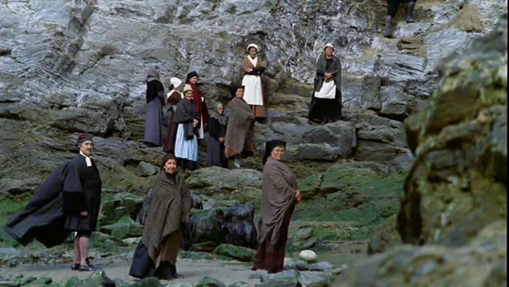 The Scarlet Letter (1973) movie still: wide — A group of people wearing 19th-century attire gather on a rocky coastal cliff. The figures are…