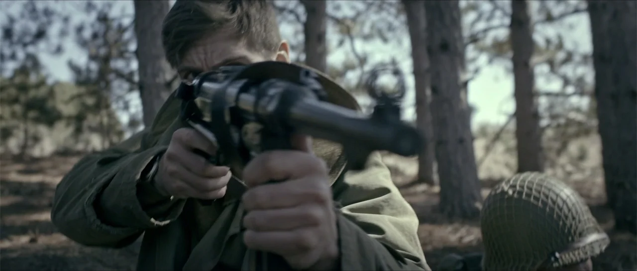 Battle of the Bulge (1965) movie still: medium — A soldier kneels within a dense woodland area during the mid-20th century. The soldier holds a…