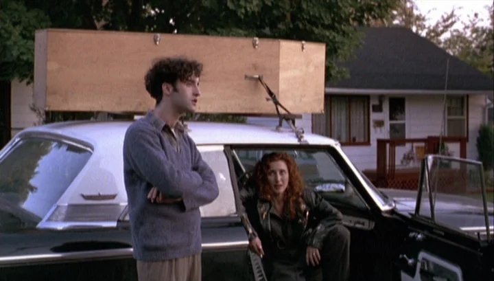 Highway 61 (1991) movie still: fullbody — A wooden coffin is strapped to the roof of a parked black sedan. A man stands beside the driver side door…