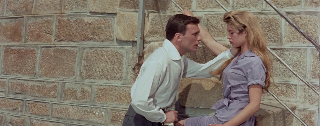 And God Created Woman (1956) movie still: medium — A man and woman stand against a large, uneven beige stone masonry wall. The man faces the woman…