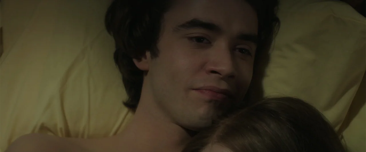 If I Stay (2014) movie still: closeup — A dimly lit bedroom features yellow bedsheets and a man lying beside another person. The man gazes forward…