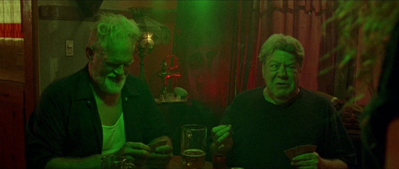 Bliss movie still: medium — A dimly lit room bathed in artificial green light featuring a card-playing area. Two older men sit at a table holding…