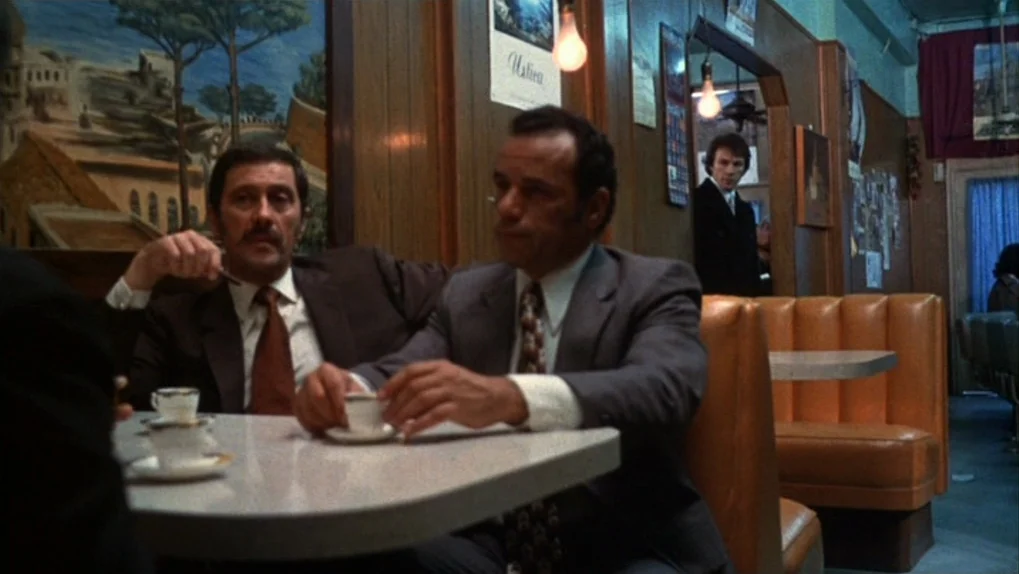 Mean Streets (1973) movie still: medium — A vintage 1970s diner with orange vinyl booths and wooden wall paneling. Two men in suits sit across from…