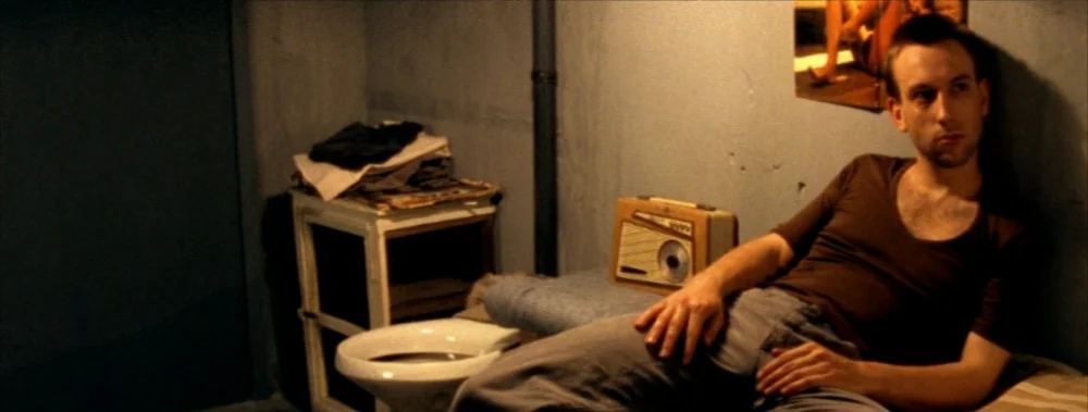 Carne (1991) movie still: medium — A dimly lit, sparse room featuring a bed, a nearby toilet, and a radio. A man lies on the bed against the wall…