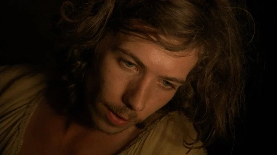 The Tudors (2007) movie still: closeup — A dark, minimally furnished interior space is cast in shadows. A young man with long hair gazes downward at…