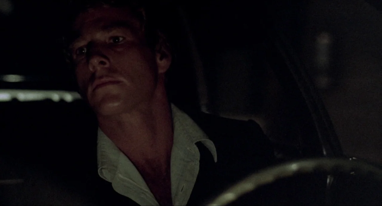 The Driver (1978) movie still: closeup — The interior of a moving vehicle is dark at night. A young man drives while illuminated by faint ambient…