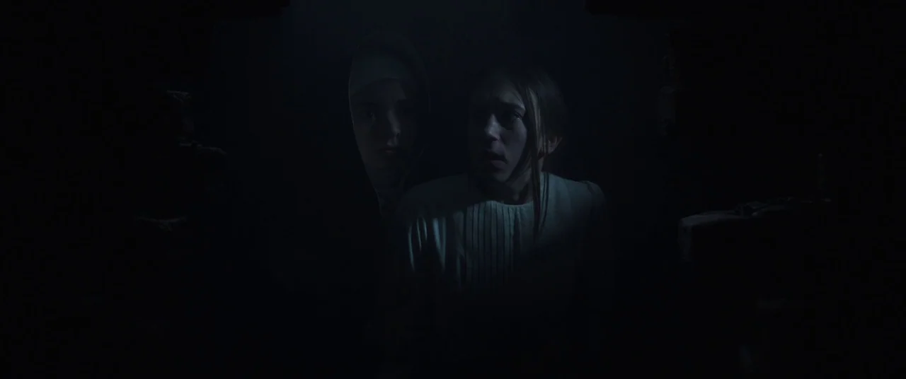 The Nun (2018) movie still: medium — Two women stand in a dark, confined space with shadowed walls surrounding them. One woman looks forward while…