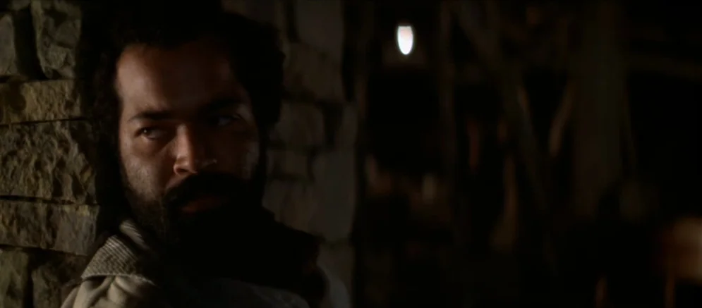 Ride With The Devil movie still: closeup — An interior space features a rough-hewn stone wall illuminated by low, dim light. A man with a full beard…