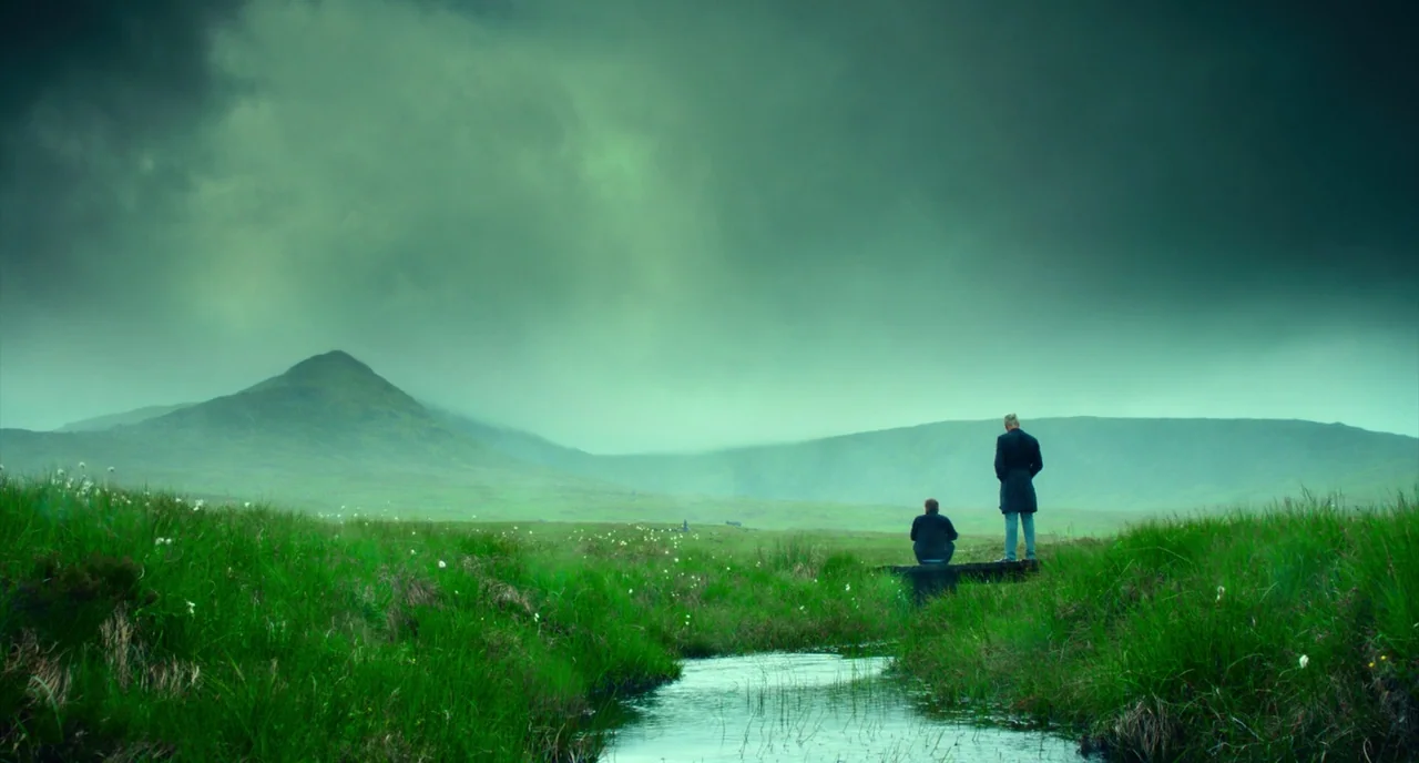 T2 Trainspotting (2017) movie still: wide — A vast, green Highland moor stretches toward a misty mountain under an overcast sky. Two men in dark…