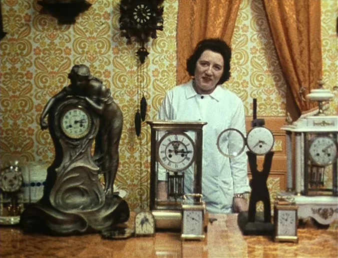 Daguerréotypes (1976) movie still: medium — A woman stands behind a table cluttered with various antique timepieces in a shop. Patterned orange and…