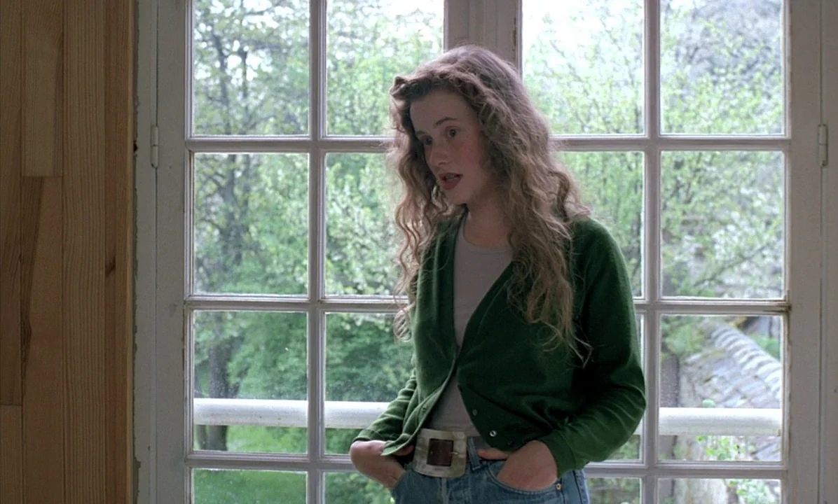 A Tale of Springtime (1990) movie still: medium — A woman stands before a large multi-pane window with lush green trees visible outside. She wears a…