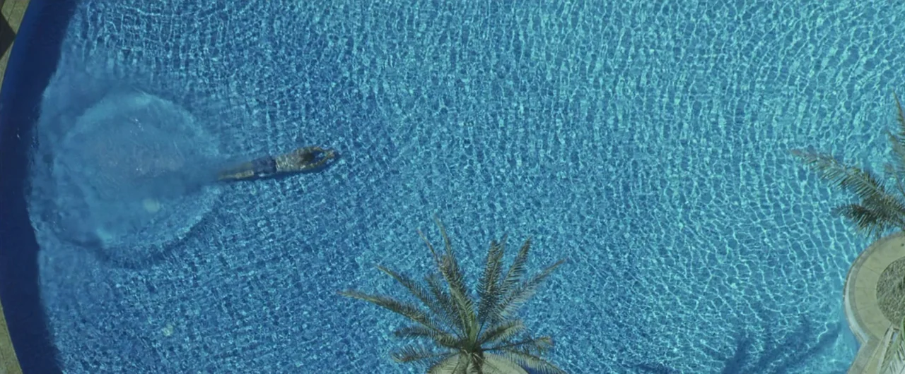 Syriana (2005) movie still: wide — A top-down view of a bright blue tiled pool with rippling water patterns. A lone person swims toward the left…