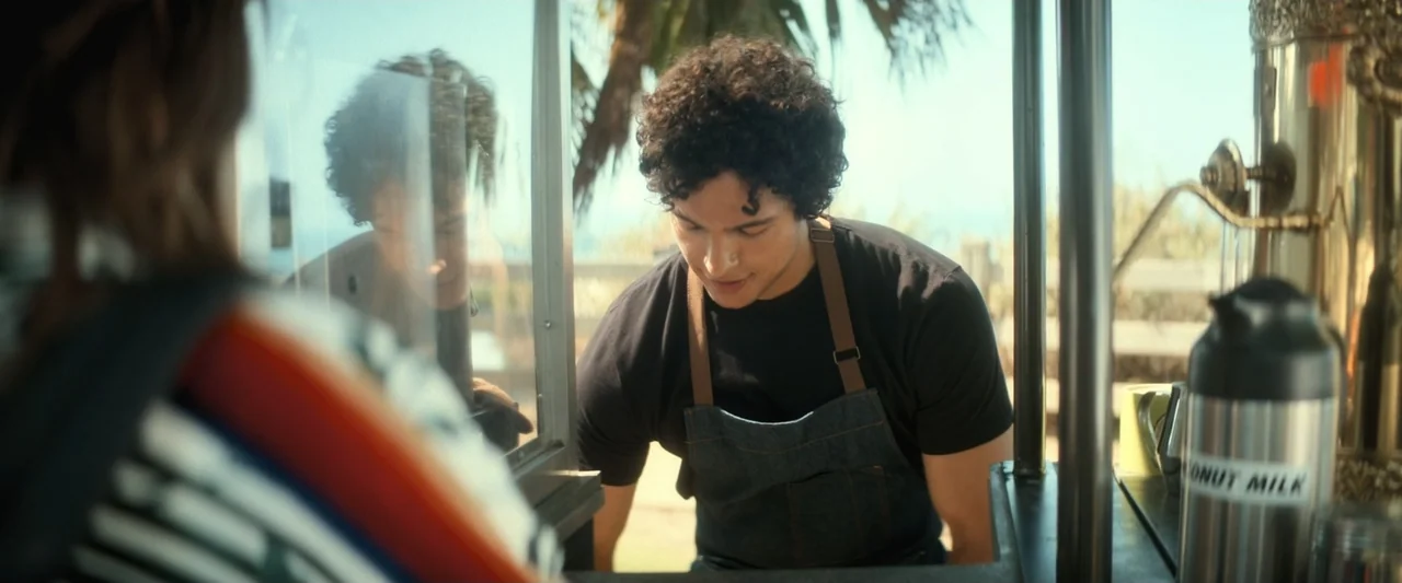 I Love America (2022) movie still: medium — An outdoor mobile beverage cart stands in a sunlit area near palm trees. A man with curly hair and a…