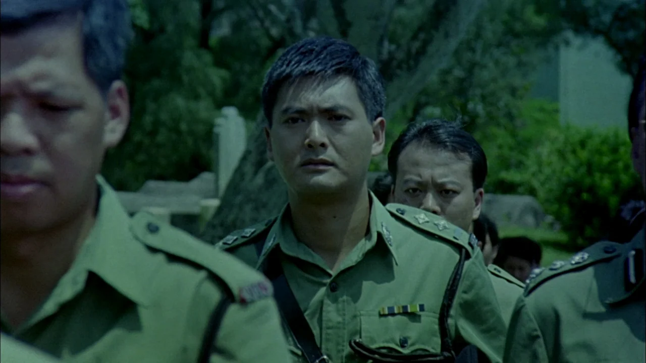 Hard Boiled (1992) movie still: medium — A group of men in olive green uniforms walk together through a park-like cemetery. The men look ahead as…