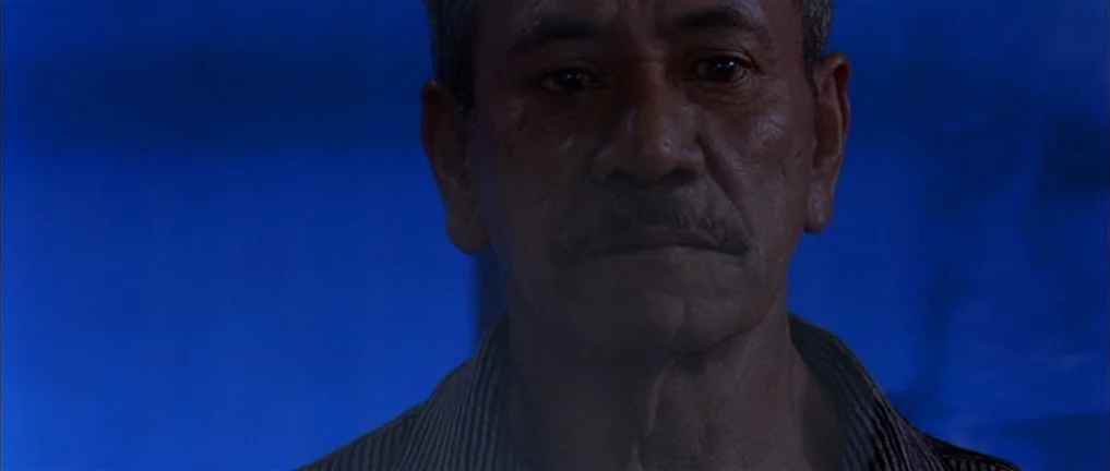 The Intruder (2004) movie still: closeup — An interior space bathed in intense blue light against a dark background. An elderly man with a thin…