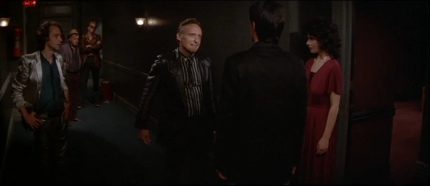 Blue Velvet (1986) movie still: fullbody — A dimly lit narrow hotel hallway with dark walls and dim sconce lighting. A small group of people stand…