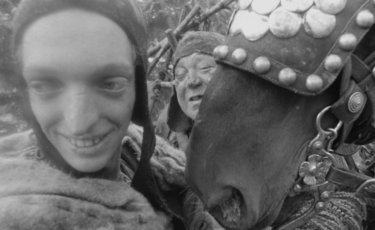 Hard To Be A God (2013) movie still: medium — An outdoor field with a wooden rack visible in the blurred background. A young person smiles closely…