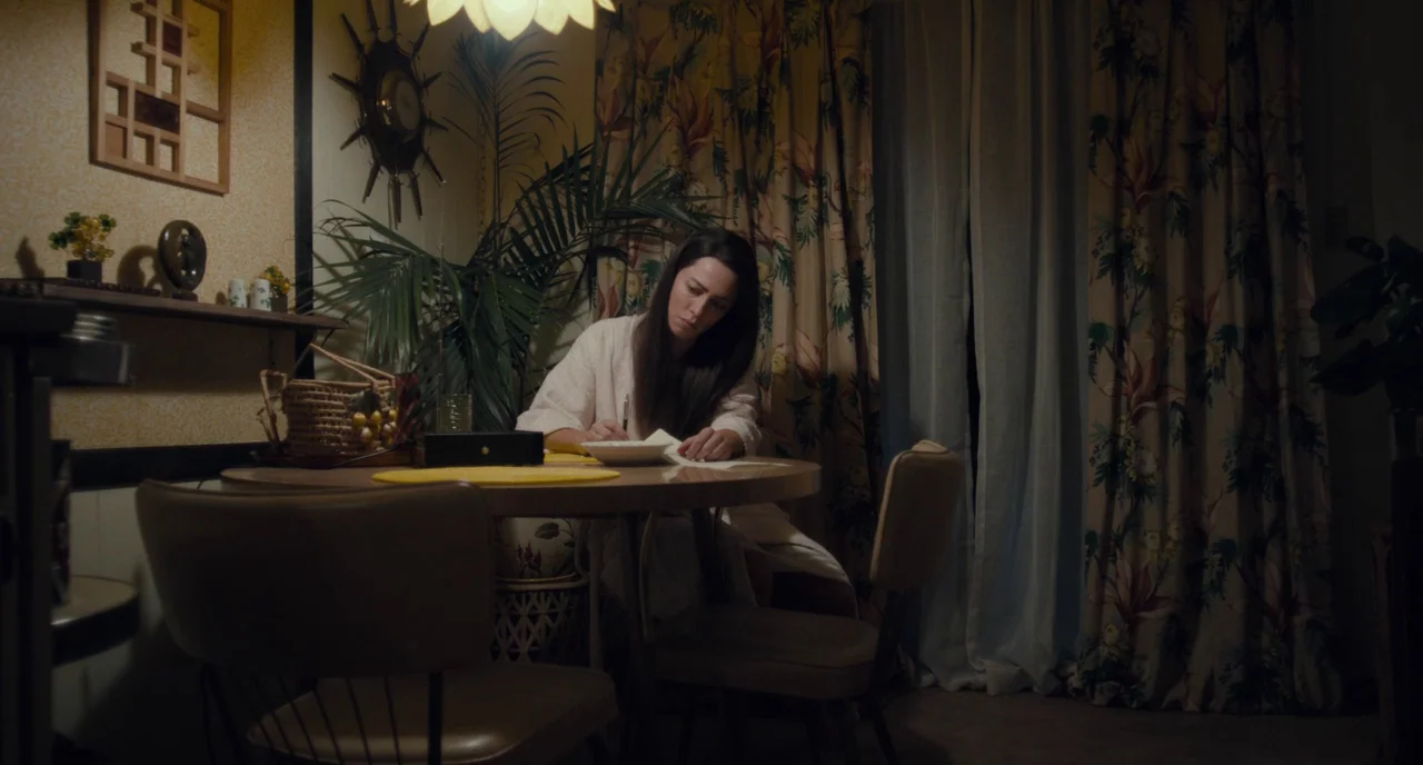 Christine (2016) movie still: medium — A 1970s styled space features floral curtains, a sunburst wall clock, and wallpaper. A woman wearing a robe…