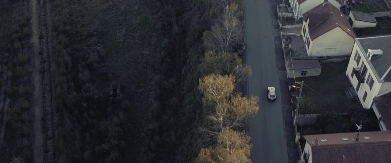 Meanwhile On Earth (2024) movie still: establishing — An aerial view shows a suburban street bordered by trees and train tracks. A single white van…