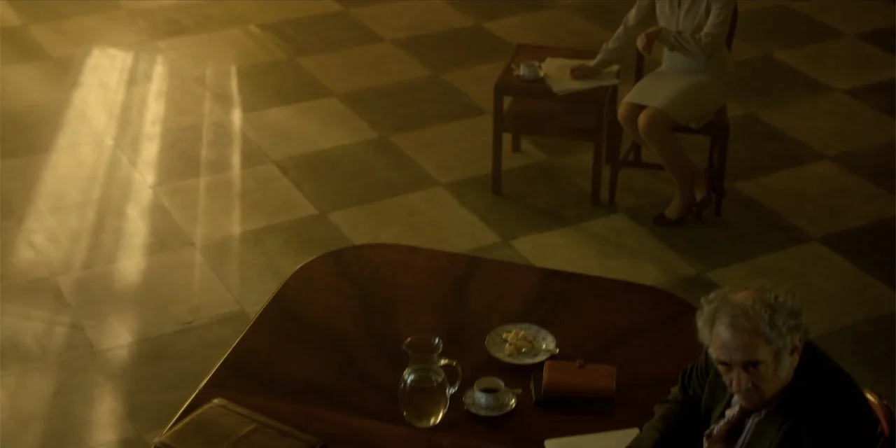 Oslo (2021) movie still: wide — A spacious room features a checkered stone floor and wooden furniture under warm light. An older man and a…